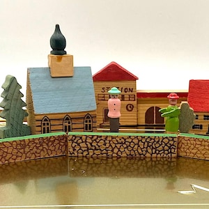 Puede incluir: Una escena de ciudad de juguete de madera con edificios, árboles y figuras. Los edificios tienen tejados rojos y azules, incluyendo una iglesia, casas y una estación. Dos torres con puntas cónicas verdes flanquean la escena.
