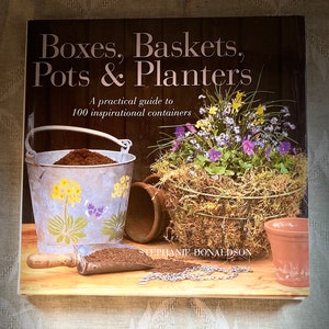Puede incluir: Un libro titulado "Boxes, Baskets, Pots & Planters: A practical guide to 100 inspirational containers" de Stephanie-Donaldson. La portada presenta una naturaleza muerta rústica con un cubo de metal, una cesta tejida, una maceta de terracota y una pala de tierra.