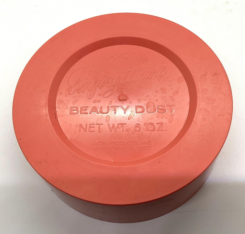 May include: A round, orange plastic container with a white label that reads "Avon Unforgettable Beauty Dust Net Wt. 6 oz. Avon Products Inc. New York, NY 10020."
