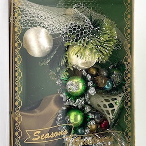 May include: A vintage Christmas ornament display in a green box. The arrangement includes green and silver baubles, faux evergreen branches, and a gold ribbon. The box has gold trim and the words "Seasons Greetings".