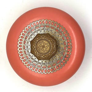 May include: A round, coral-colored container with a gold-colored, decorative lid. The lid features a circular, filigree design with a smaller, octagonal, gold-colored design in the center.