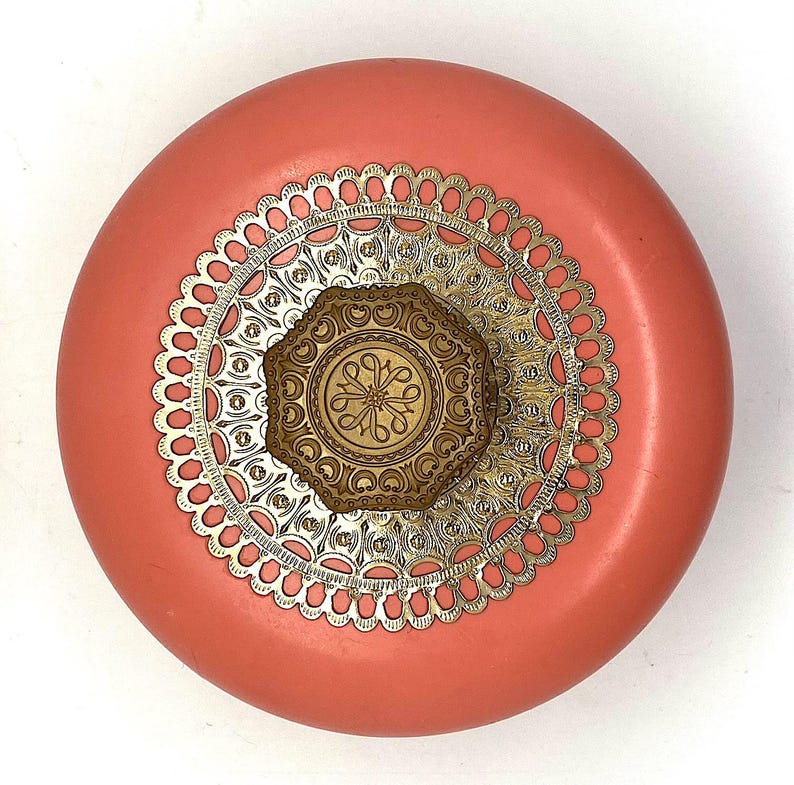 May include: A round, coral-colored container with a gold-colored, decorative lid. The lid features a circular, filigree design with a smaller, octagonal, gold-colored design in the center.