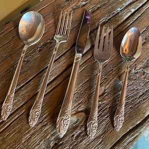 May include: Five pieces of silverware with ornate floral designs on the handles. The set includes a butter knife, a dinner fork, a teaspoon, and two dinner forks.