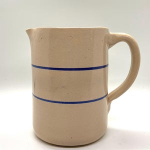 以下が含まれることがあります： ベージュ色のセラミックピッチャー。曲線的なハンドルと注ぎ口が付いています。2本の水平な青いストライプがピッチャーの胴体を囲んでいます。クラシックで素朴なデザインです。