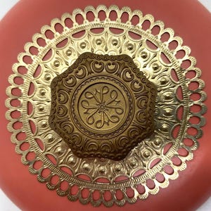 May include: A coral pink round box with a gold metal lid. The lid has a decorative design with a central octagonal medallion. The medallion has a floral design with a stylized letter "L" in the center.
