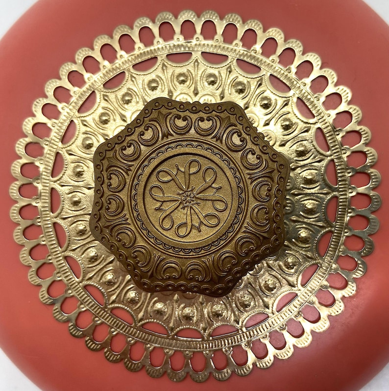 May include: A coral pink round box with a gold metal lid. The lid has a decorative design with a central octagonal medallion. The medallion has a floral design with a stylized letter "L" in the center.