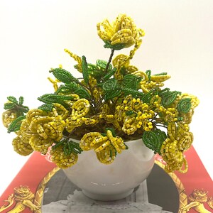 May include: A beaded floral arrangement featuring yellow flowers and green leaves. The flowers are in a white ceramic pot. The arrangement is set against a white background.