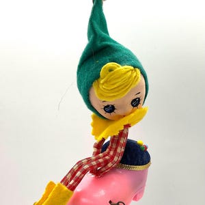 May include: A vintage Christmas ornament featuring a doll with a green felt hat, yellow hair, and a yellow collar. The doll is seated on a pink shoe with red and white checkered legs and yellow boots. The ornament is suspended by a gold wire.