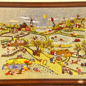 May include: Embroidered landscape scene in a wooden frame. The artwork depicts a detailed village with houses, farms, a lighthouse, and people engaged in various activities. The color palette includes greens, yellows, reds, and blues, creating a vibrant and charming aesthetic.