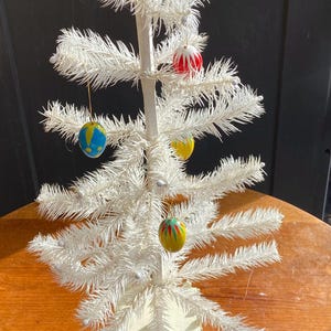 May include: A small, white artificial Christmas tree with colourful baubles. The tree has a rectangular base and is decorated with red, yellow, and blue ornaments. The tree is set on a wooden table.