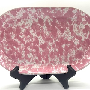 May include: Oval serving platter with a pink and white speckled pattern. The ceramic dish is displayed on a black stand. The platter has a raised rim and a smooth surface.