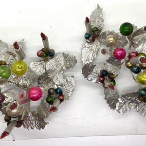 May include: Two silver candle holders with a Christmas theme. Each holder features a silver base with a cluster of green, red and gold leaves and ornaments. The holders are decorated with red candles.