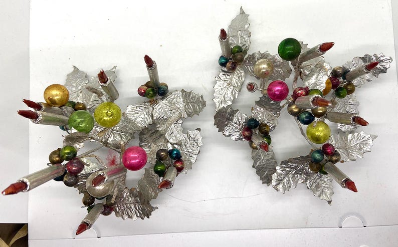 May include: Two silver candle holders with a Christmas theme. Each holder features a silver base with a cluster of green, red and gold leaves and ornaments. The holders are decorated with red candles.