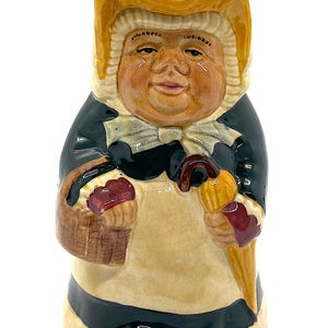 Könnte beinhalten: Eine Keramikfigur einer Frau mit einem gelben Hut, einem grünen Kleid und einem Korb und einem Regenschirm in der Hand. Die Figur ist handbemalt mit Details in Braun, Rot und Gelb.