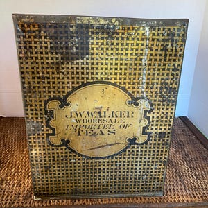 Puede incluir: Lata de té vintage de metal con un patrón tejido negro y dorado. La lata tiene una etiqueta dorada con el texto "J.W. WALKER WHOLESALE IMPORTER OF TEAS".