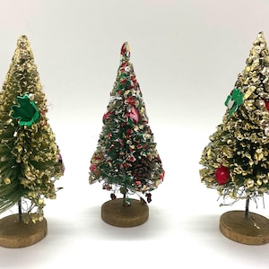 May include: Three miniature Christmas trees with gold, green, and red decorations. Each tree stands on a small wooden base and is adorned with ornaments. These trees are a festive holiday decoration.