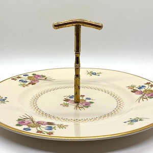 Puede incluir: Una bandeja de servir de cerámica blanca con un borde dorado y un patrón floral. La bandeja tiene un asa de metal dorado.