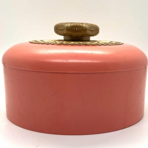 May include: A pink plastic powder jar with a gold-colored lid and a decorative knob. The knob has a gold-colored design.