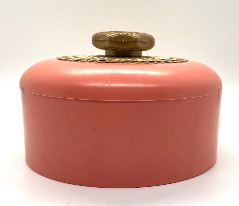 May include: A pink plastic powder jar with a gold-colored lid and a decorative knob. The knob has a gold-colored design.