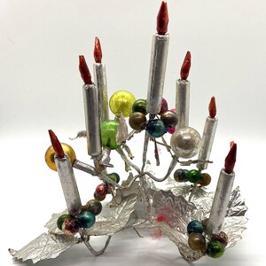 May include: A silver metal Christmas tree ornament featuring seven silver candles with red candle flames and colourful glass beads. The base of the ornament is made of silver foil and resembles leaves.