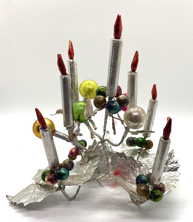 May include: A silver metal Christmas tree ornament featuring seven silver candles with red candle flames and colourful glass beads. The base of the ornament is made of silver foil and resembles leaves.