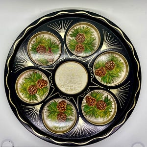 May include: A black metal serving tray with a gold rim and six individual serving sections. Each section features a painted design of pine branches and pine cones. The centre of the tray has a gold inscription.