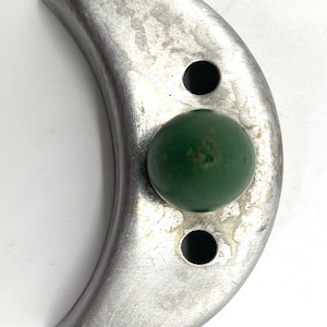 May include: A silver-toned, crescent-shaped object with a green, spherical accent. Two circular holes are visible on the crescent. The object has a brushed metal finish and a slightly worn appearance. The green sphere is centered on the crescent.