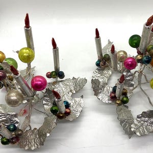 May include: Two silver candle holders with a holly leaf design. Each holder has six silver candles with red tips and is decorated with colourful glass baubles.