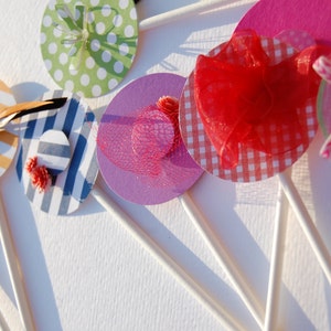 May include: Six colorful paper cupcake toppers with different designs, including stripes, polka dots, and gingham patterns. Each topper is attached to a white stick.