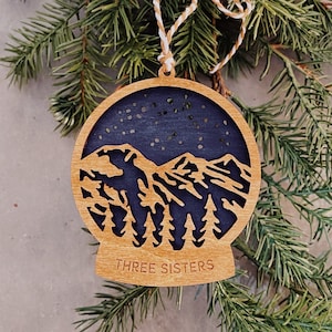 May include: Wooden Christmas ornament featuring a silhouette of the Three Sisters mountains with a starry night sky. The ornament is brown with a blue sky and is labeled "THREE SISTERS".