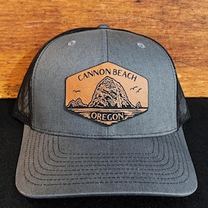 May include: A gray and black trucker hat with a brown leather patch that says "Cannon Beach, Oregon" and features a graphic of Haystack Rock.