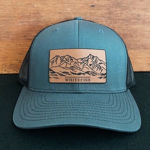 May include: A dark gray trucker hat with a brown leather patch featuring a mountain range and the text "Whitefish".
