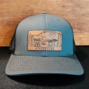 May include: A gray and black trucker hat with a wooden patch featuring a mountain and tree design. The patch reads "WINTER PARK" in a serif font. The hat has a curved brim and a mesh back.