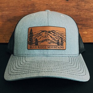 May include: A gray and black trucker hat with a brown leather patch featuring a mountain range and the text "BLUE RIDGE MOUNTAINS".