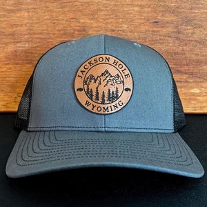 May include: A gray and black trucker hat with a brown leather patch that says "Jackson Hole Wyoming" and features a mountain range and pine trees.