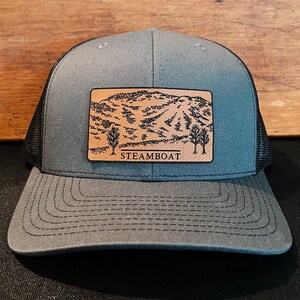 May include: A gray and black trucker hat with a brown leather patch featuring a mountain scene and the text "STEAMBOAT".