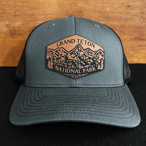 May include: A dark gray trucker hat with a brown leather patch that says "Grand Teton National Park" with a mountain range design.