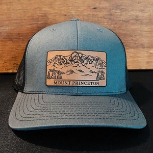 May include: A gray and black trucker hat with a brown leather patch featuring a mountain range and the text "MOUNT PRINCETON".