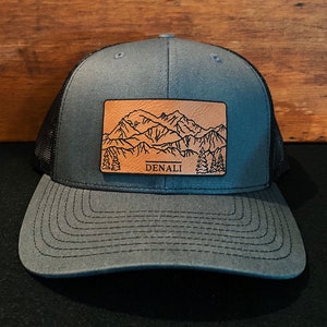 May include: A gray and black trucker hat with a brown leather patch featuring a mountain range and the word "DENALI" in black.
