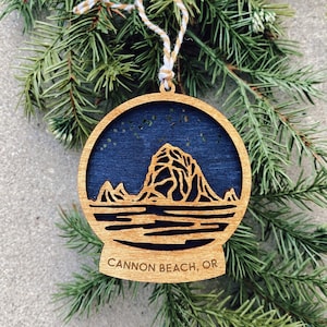 May include: Wooden Christmas ornament with a blue and white design of Cannon Beach, Oregon. The ornament is round and has a string for hanging.