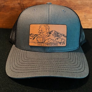May include: A gray and black trucker hat with a brown leather patch featuring a drawing of a geyser and the text "YELLOWSTONE".