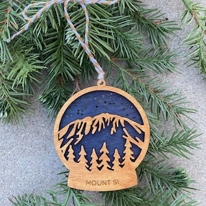 May include: Wooden ornament shaped like a snow globe with a blue mountain silhouette and a forest of trees at the base. The ornament is decorated with white stars and the text "MOUNT SI" is printed on the bottom.