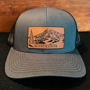 May include: A gray and black trucker hat with a brown leather patch featuring a mountain silhouette and the text "MOUNT RAINIER".