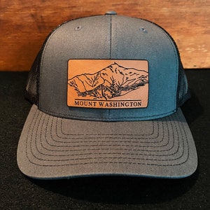 May include: A gray and black trucker hat with a brown leather patch featuring a mountain range and the text "MOUNT WASHINGTON".