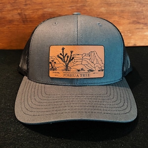 May include: A gray and black trucker hat with a brown leather patch featuring a Joshua Tree silhouette and the text "Joshua Tree".