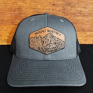 May include: A gray and black trucker hat with a brown leather patch featuring a mountain range and the text "Mount Whitney California".
