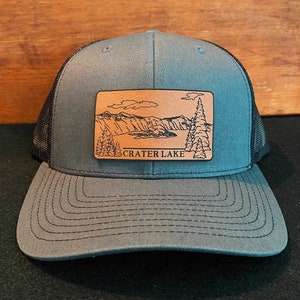 Peut inclure: Un chapeau de camionneur gris et noir avec un patch en cuir brun représentant un dessin du lac Crater et le texte "CRATER LAKE".