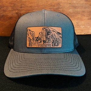 May include: A gray and black trucker hat with a brown leather patch featuring a black and white illustration of Yosemite National Park and the word "Yosemite".