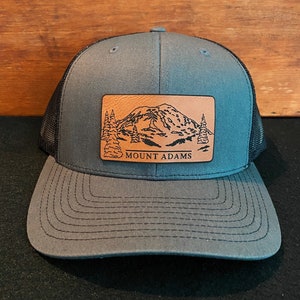May include: A gray and black trucker hat with a brown leather patch featuring a mountain scene and the text "MOUNT ADAMS".