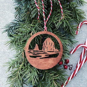 May include: A wooden Christmas ornament featuring a silhouette of Haystack Rock, a famous landmark in Oregon. The ornament is brown and has a red and white twine hanger.